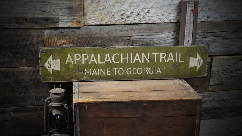 May include: A rectangular olive green wooden sign with distressed edges featuring the white text APPALACHIAN TRAIL and MAINE TO GEORGIA. The sign includes white directional arrows on both ends, with an S on the left and an N on the right.