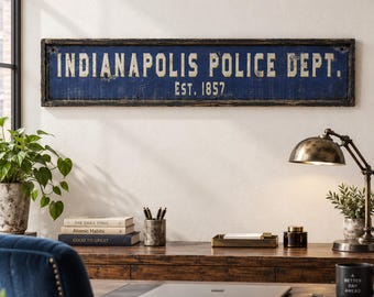 May include: A rectangular wall sign with a distressed dark blue finish and a black frame. The text in white, weathered lettering reads INDIANAPOLIS POLICE DEPT. EST. 1857. The sign has a horizontal orientation and a worn, aged appearance.