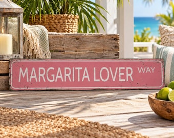 May include: A rectangular metal street sign with a distressed pink finish and a white border. The sign features the white text MARGARITA LOVER WAY in a bold, sans-serif font. The surface shows signs of wear with small dark speckles throughout.