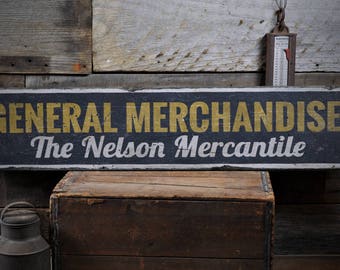 Op de afbeelding: Een zwart-wit houten bord met een gouden pijl die naar rechts wijst. Het bord heeft de tekst "General Merchandise The Nelson Mercantile".