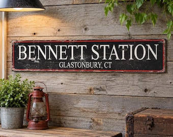 May include: A rectangular black sign with white lettering that reads BENNETT STATION and GLASTONBURY, CT. The sign features thin red horizontal lines along the top and bottom edges and has a distressed finish with visible wear and texture.