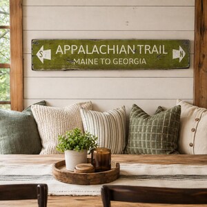 May include: A rectangular wooden sign with a distressed green finish and white text reading APPALACHIAN TRAIL MAINE TO GEORGIA. The sign features white directional arrows on both ends, with an S on the left and an N on the right side.