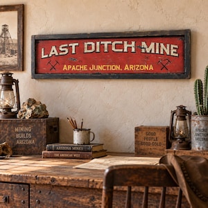 May include: A rectangular wooden sign with a distressed red background and a dark frame. The text reads LAST DITCH MINE in large white letters, with APACHE JUNCTION, ARIZONA in yellow below. Two black crossed pickaxe icons flank the text.