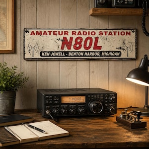 May include: A rectangular metal sign with a distressed white finish, featuring the red text AMATEUR RADIO STATION, the call sign N80L in large red letters, and the black text KEN JEWELL - BENTON HARBOR, MICHIGAN, flanked by two black radio tower icons.
