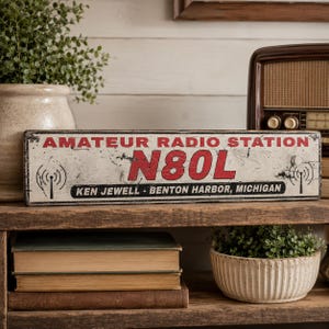 May include: A rectangular metal sign with a distressed white finish, featuring the red text AMATEUR RADIO STATION and N80L. Below, black text reads KEN JEWELL - BENTON HARBOR, MICHIGAN. The sign includes black radio tower icons on both sides.