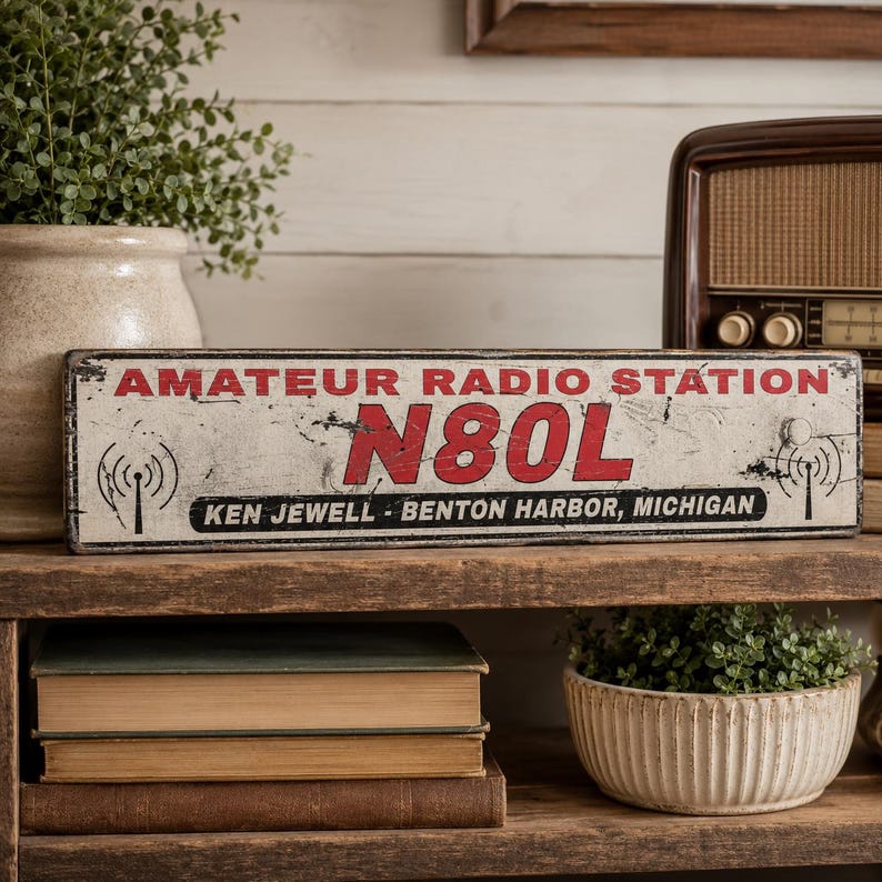 May include: A rectangular metal sign with a distressed white finish, featuring the red text AMATEUR RADIO STATION and N80L. Below, black text reads KEN JEWELL - BENTON HARBOR, MICHIGAN. The sign includes black radio tower icons on both sides.
