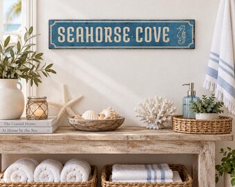 May include: A rectangular blue wooden sign with distressed edges featuring the white text SEAHORSE COVE and a seahorse illustration. The sign has a decorative border and is mounted on a wall above a wooden console table with various decor items.