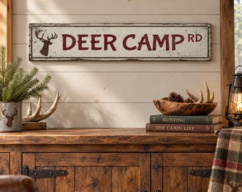 May include: A rectangular wall sign with a distressed white finish and a dark red border. The sign features a dark red silhouette of a deer head on the left and the text DEER CAMP RD in bold, dark red capital letters across the center.