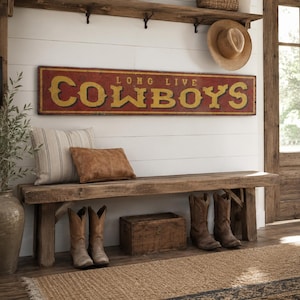 May include: A rectangular wooden sign with a distressed red background and yellow lettering that reads LONG LIVE COWBOYS. The sign is mounted on a white horizontal plank wall above a wooden bench, a striped pillow, a brown pillow, and boots.