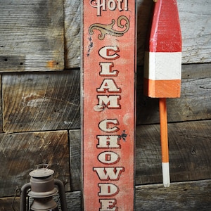 Puede incluir: Un letrero rústico y desgastado con las palabras "Hot! Clam Chowder" en letras blancas. Una boya roja, blanca y naranja cuelga de una cuerda. Una linterna vintage con lente de cristal rojo se encuentra sobre una superficie de madera.
