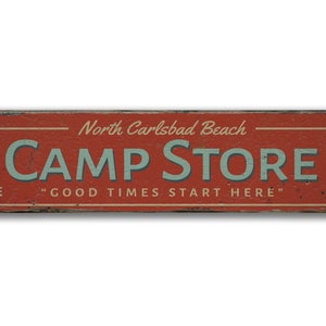 May include: Red wooden sign with white text that reads "North Carlsbad Beach CAMP STORE" and "GOOD TIMES START HERE".  The sign also lists items for sale: "ICE, BEER, BAIT, TACKLE".  A hand pointing right with the text "1/2 Mi." is on the right side of the sign.