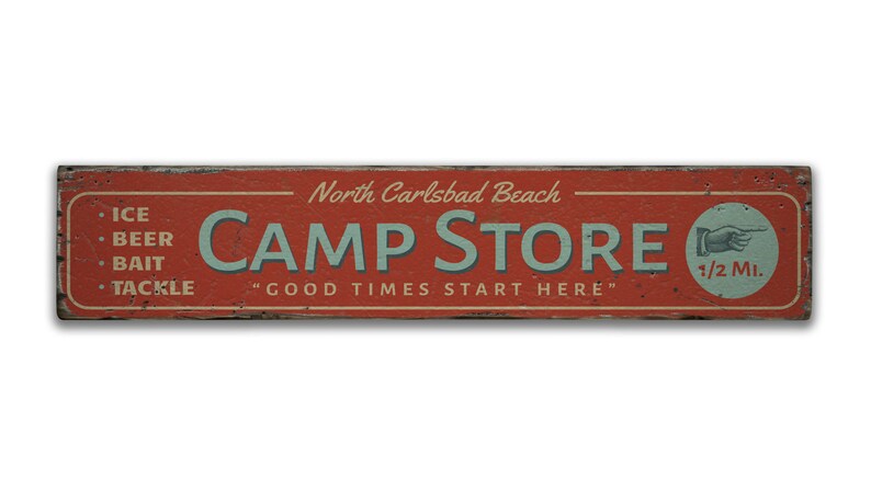 May include: Red wooden sign with white text that reads "North Carlsbad Beach CAMP STORE" and "GOOD TIMES START HERE".  The sign also lists items for sale: "ICE, BEER, BAIT, TACKLE".  A hand pointing right with the text "1/2 Mi." is on the right side of the sign.