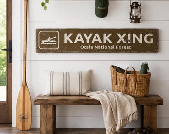 Könnte beinhalten: Ein rechteckiges braunes Holzschild mit weißem Text KAYAK XING und Ocala National Forest sowie einer weißen Grafik einer Person im Kajak. Das Schild ist an einer weißen Wand über einer Holzbank mit Kissen und einem geflochtenen Korb montiert.