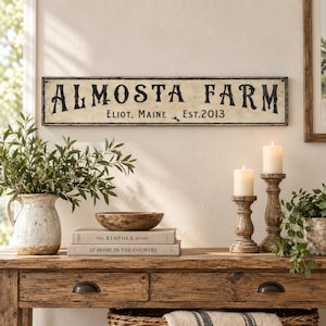 May include: A rectangular wall sign with a distressed cream background and black lettering that reads ALMOSTA FARM, ELIOT, MAINE, EST. 2013. The sign features a thin black border and a weathered finish, mounted horizontally on a light-colored wall.
