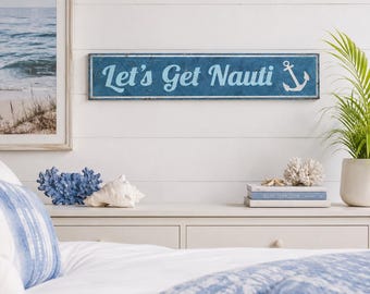 May include: A rectangular blue wooden sign with a distressed finish featuring the white text Let's Get Nauti and a white anchor graphic on the right side. The sign has a thin white border and is mounted horizontally on a white shiplap wall.