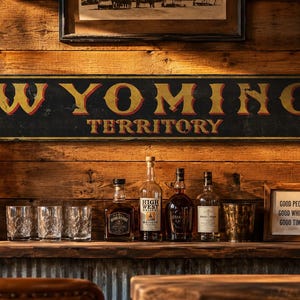 May include: A rectangular black sign with yellow and red lettering that reads WYOMING TERRITORY. The sign is mounted on a horizontal wood plank wall above a bar area featuring several glass bottles, three clear glasses, and a small framed sign.