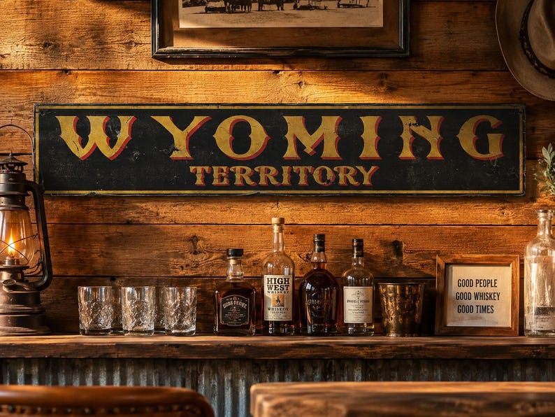 May include: A rectangular black sign with yellow and red lettering that reads WYOMING TERRITORY. The sign is mounted on a horizontal wood plank wall above a bar area featuring several glass bottles, three clear glasses, and a small framed sign.