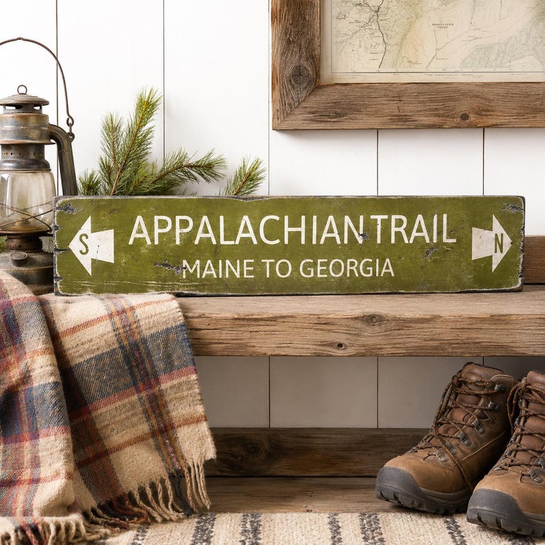 May include: A rectangular olive green wooden sign with distressed edges featuring the white text APPALACHIAN TRAIL and MAINE TO GEORGIA. The sign includes white directional arrows on both ends, with an S on the left and an N on the right.