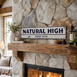 May include: A rectangular metal sign with a distressed white finish and dark blue lettering that reads NATURAL HIGH and VRBO# 34490. The sign features a thin dark blue border and a weathered, aged appearance with visible surface imperfections.