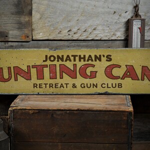 Könnte beinhalten: Ein gelbes Holzschild mit roten Buchstaben, auf dem "Jonathan's Hunting Camp Retreat & Gun Club" steht.