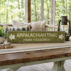 May include: A rectangular olive green wooden sign with white text reading APPALACHIAN TRAIL and MAINE TO GEORGIA. The sign features white directional arrows on both ends, with an S on the left arrow and an N on the right arrow, indicating directions.
