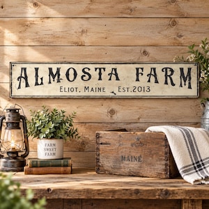 May include: A rectangular off-white metal sign with black distressed lettering reading ALMOSTA FARM, ELIOT, MAINE, EST. 2013. The sign features a weathered finish and is displayed horizontally against a light brown horizontal wood plank wall.