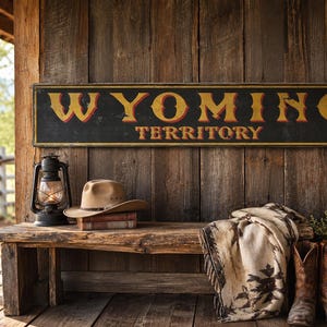May include: A rectangular black sign with yellow and red lettering that reads WYOMING TERRITORY. Below the sign sits a tan cowboy hat on books, a black lantern, a patterned beige blanket, and a pair of brown leather cowboy boots on a bench.