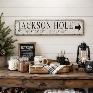 May include: A rectangular wooden sign with a distressed white finish features the black text JACKSON HOLE, an arrow pointing right, and coordinates N 43&deg; 28' 47" 110&deg; 45' 44". The sign is mounted on a white horizontal plank wall above a wooden table.