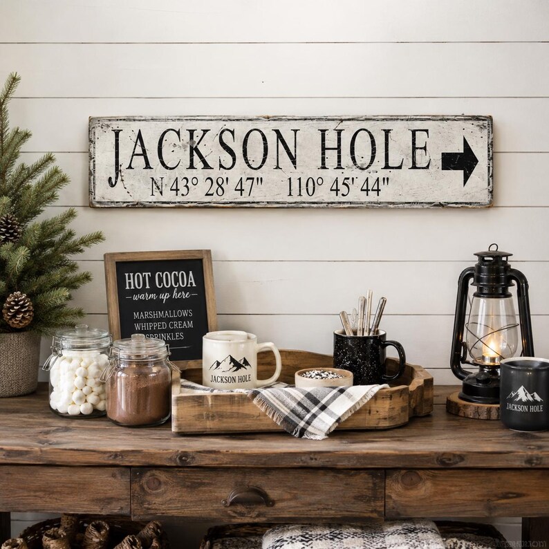 May include: A rectangular wooden sign with a distressed white finish features the black text JACKSON HOLE, an arrow pointing right, and coordinates N 43&deg; 28' 47" 110&deg; 45' 44". The sign is mounted on a white horizontal plank wall above a wooden table.