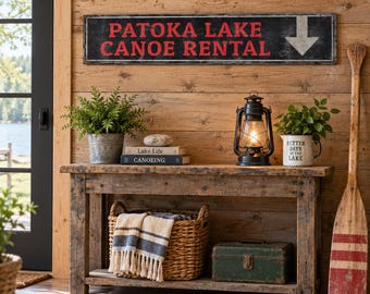 Könnte beinhalten: Ein rechteckiges schwarzes Schild mit roter Aufschrift PATOKA LAKE CANOE RENTAL und einem weißen Pfeil nach unten. Das Schild ist an einer hellbraunen Holzwand über einem rustikalen Holzkonsolentisch mit Laterne und Korb befestigt.