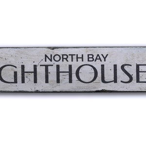 May include: A white wooden sign with black lettering that reads "North Bay Lighthouse" with a black arrow pointing to the right.