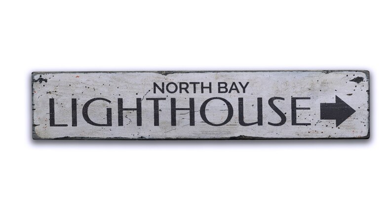May include: A white wooden sign with black lettering that reads "North Bay Lighthouse" with a black arrow pointing to the right.