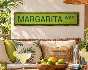 May include: A rectangular lime green metal sign with dark green text that reads MARGARITA WAY. The sign features a distressed finish with visible wear along the edges and is mounted on a textured off-white wall above a patio seating area.