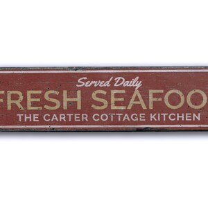 May include: A red wooden sign with white and black trim, advertising "Fresh Seafood" served daily at "The Carter Cottage Kitchen".