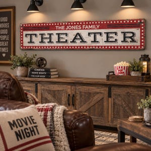 May include: A rectangular wall sign with a distressed red and white finish featuring the text THE JONES FAMILY THEATER in black lettering. The sign is bordered by a white dotted pattern and includes horizontal black lines above the main text.