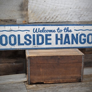 May include: A white wooden sign with blue text and wavy lines that reads "Welcome to the POOLSIDE HANGOUT". The sign is sitting on top of a wooden crate.