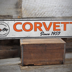 May include: A white metal sign with orange text that reads "Corvette Since 1953". The sign also features a black and white Chevrolet Corvette logo.