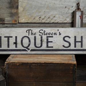 Puede incluir: Un letrero de madera blanco con letras negras que dice "The Steven's Antique Shop". El letrero tiene una flecha negra que apunta hacia la derecha.