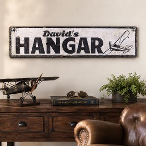 May include: A rectangular wall sign with a distressed white finish and black lettering that reads David's HANGAR. The sign features a black line-art illustration of a small aeroplane on the right side and has a dark, weathered border.