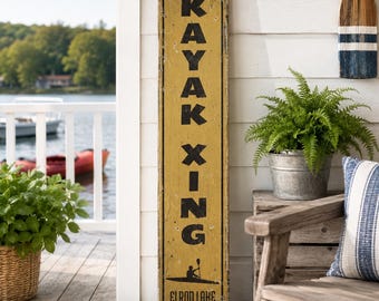 Könnte beinhalten: Ein hohes, rechteckiges, verwittertes senfgelbes Holzschild mit schwarzem Rand. Der Text "KAYAK XING" ist in fetten, schwarzen, serifenlosen Großbuchstaben gedruckt, gefolgt von einer schwarzen Silhouette eines Kajakfahrers und dem Text "ELROD LAKE".