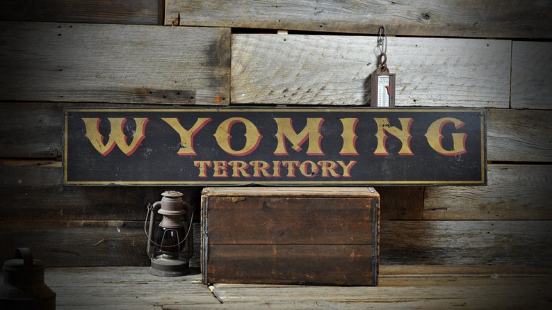 May include: A black wooden sign with gold and red lettering that reads "WYOMING TERRITORY". The sign is hanging on a rustic wooden wall.