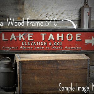Op de afbeelding: Een rood en wit bord met zwarte letters met de tekst "LAKE TAHOE ELEVATION 6,225 Largest Alpine Lake in North America". Het bord heeft een zwarte lijst en een witte pijl die naar rechts wijst. Het bord staat op een houten achtergrond. Voeg een optionele houten lijst toe voor &euro;40.