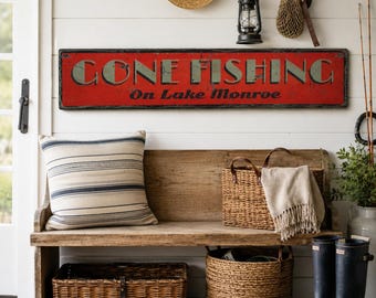 Puede incluir: Un letrero rectangular rojo con bordes desgastados que presenta el texto GONE FISHING en letras verdes grandes y On Lake Monroe en una escritura negra más pequeña. El letrero está montado en una pared blanca sobre un banco de madera.