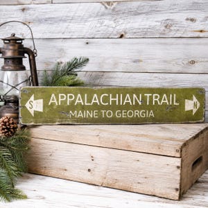 May include: A rectangular green wooden sign with distressed edges featuring the white text APPALACHIAN TRAIL and MAINE TO GEORGIA. The sign includes white arrow icons on both ends, with an S on the left arrow and an N on the right arrow.