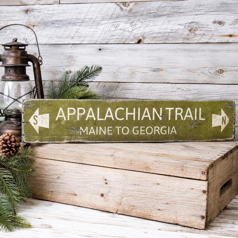 May include: A rectangular green wooden sign with distressed edges featuring the white text APPALACHIAN TRAIL and MAINE TO GEORGIA. The sign includes white arrow icons on both ends, with an S on the left arrow and an N on the right arrow.
