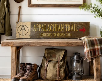 May include: A rectangular wooden sign with a dark brown finish featuring the text APPALACHIAN TRAIL and GEORGIA TO MAINE 2014 in yellow lettering. The sign includes a black circular AT logo on the left and a small red arrow on the right side.