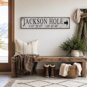 May include: A rectangular white wooden sign with a distressed finish featuring the black text JACKSON HOLE, the coordinates N 43&deg; 28' 47" 110&deg; 45' 44", and a black arrow pointing to the right, mounted on a wall above a wooden bench.