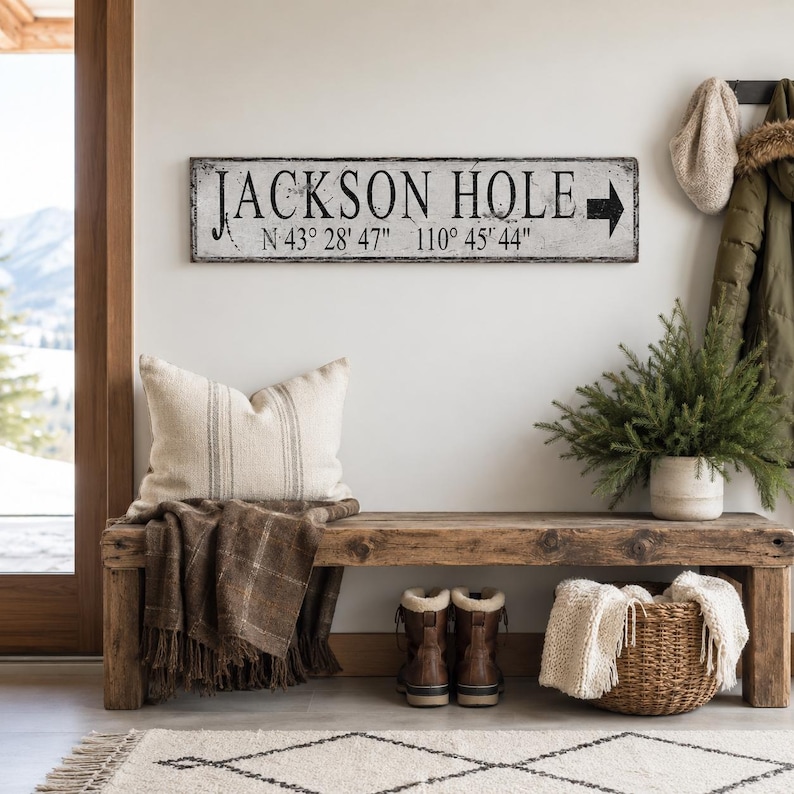 May include: A rectangular white wooden sign with a distressed finish featuring the black text JACKSON HOLE, the coordinates N 43&deg; 28' 47" 110&deg; 45' 44", and a black arrow pointing to the right, mounted on a wall above a wooden bench.