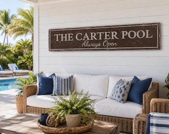 Puede incluir: Un letrero rectangular de madera marrón oscuro con letras blancas que dice THE CARTER POOL Always Open. El letrero presenta un acabado desgastado y un borde blanco fino, montado horizontalmente en una pared de listones blancos sobre un sofá.