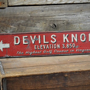 May include: Red wooden sign with white text that reads "Devils Knob Elevation 3,850 The Highest Golf Course in Virginia". A white arrow points to the left.